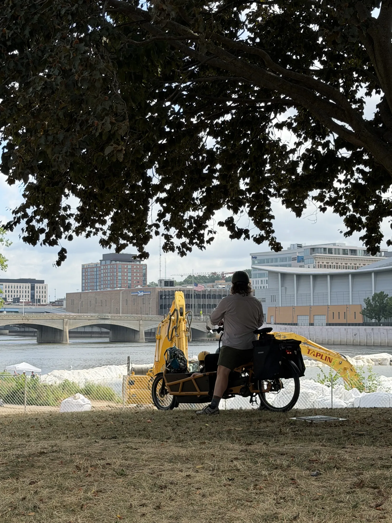 A longtail cargo e-bike stopped under a broad shade tree on a riverbank, its rider standing astride it and looking across the water at a yellow excavator working among white stone, with a bridge and downtown buildings behind